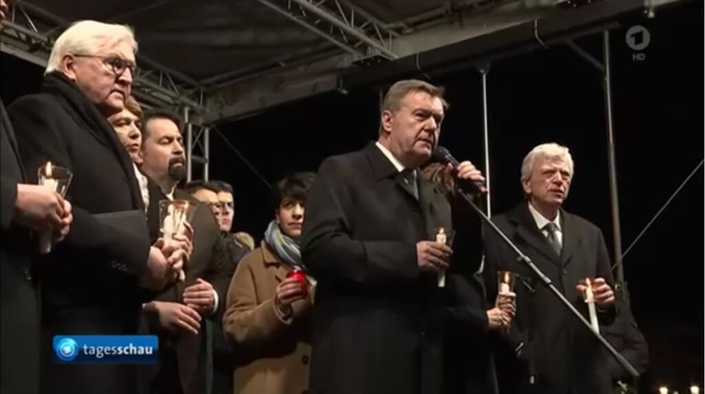 Politiker:innen und andere Sprecher:innen auf Bühne halten Kerzen in ihren Händen